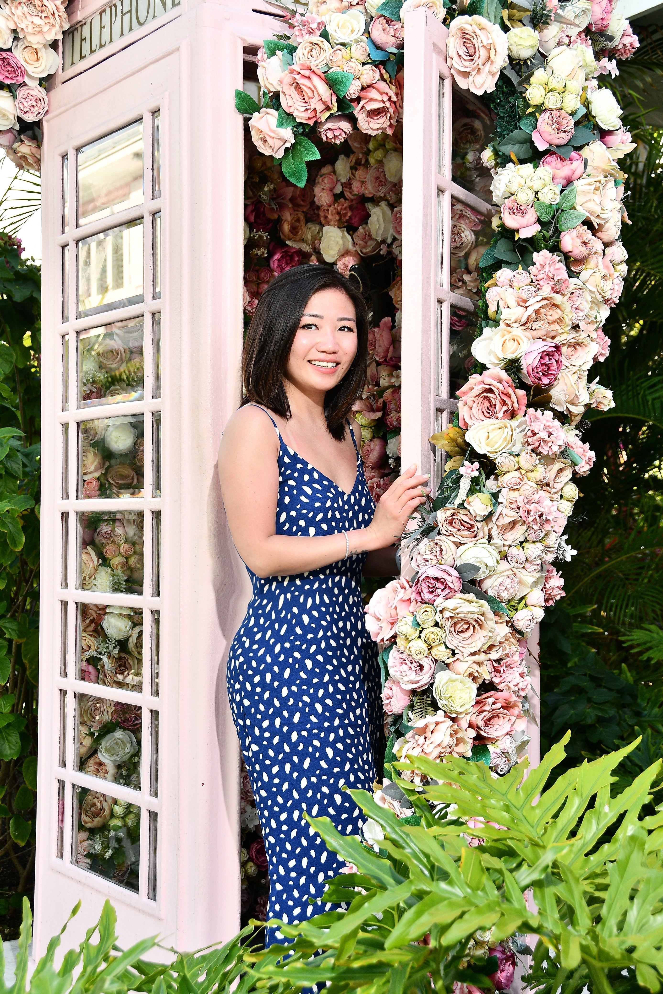 Woman owner of Marina Floral  wearing a blue summer dress standing in a floral archway with a vintage phone booth.